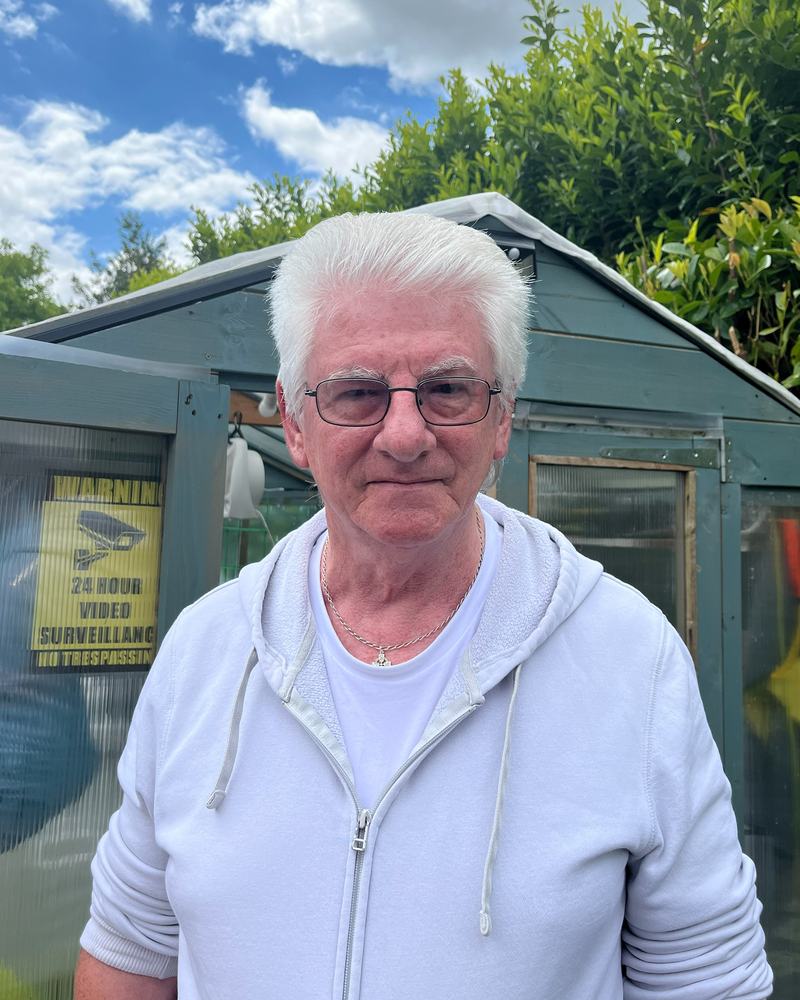 A close up of a man with white hair and glasses, in a light blue jacket, stood outside in a garden.