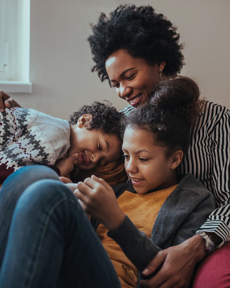 A woman and two children, a boy and girl are sat cuddling and smiling on a light brown sofa.