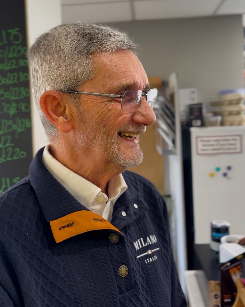 A man with light grey hair and a blue jacket smiling, white kitchen appliances are in the background.