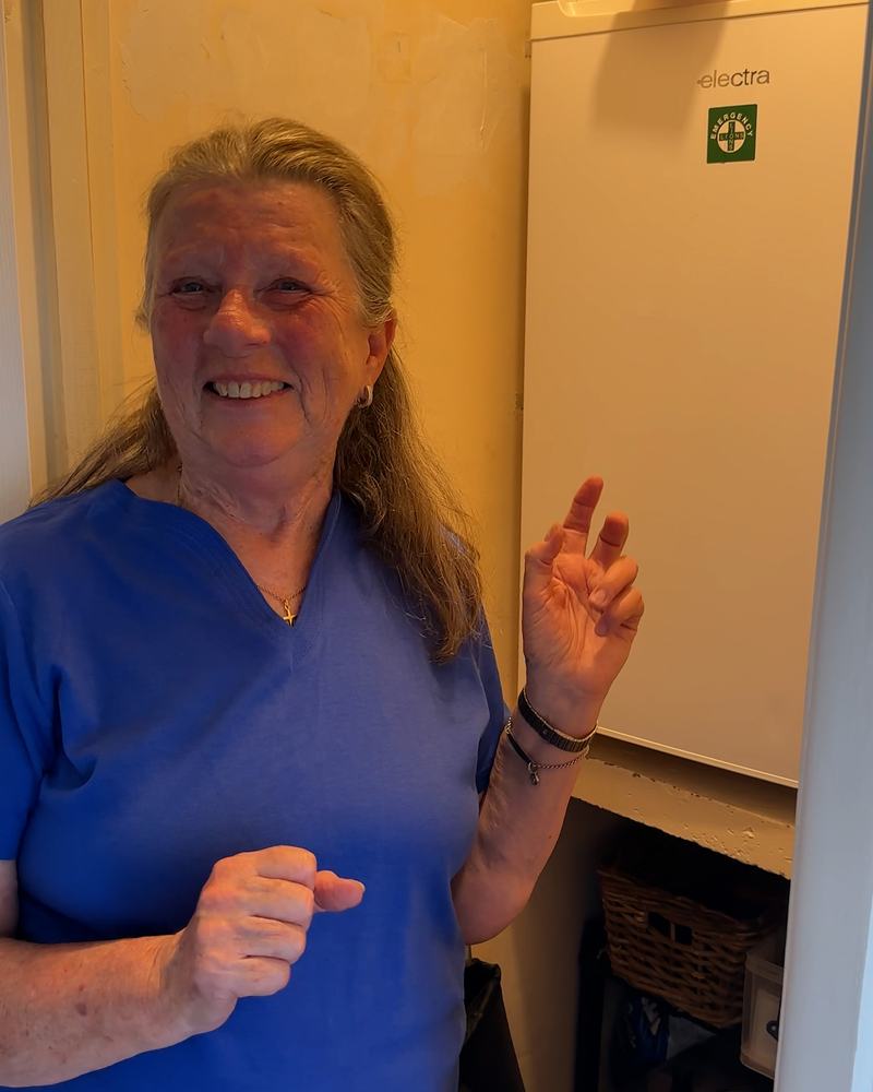 A woman wearing a blue shirt is smiling and gesturing with their hands in a room with a white appliance and various stored items.