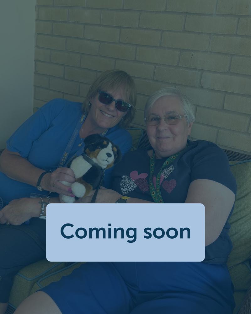 Two women are smiling and sat down together on a sofa hold a toy dog.