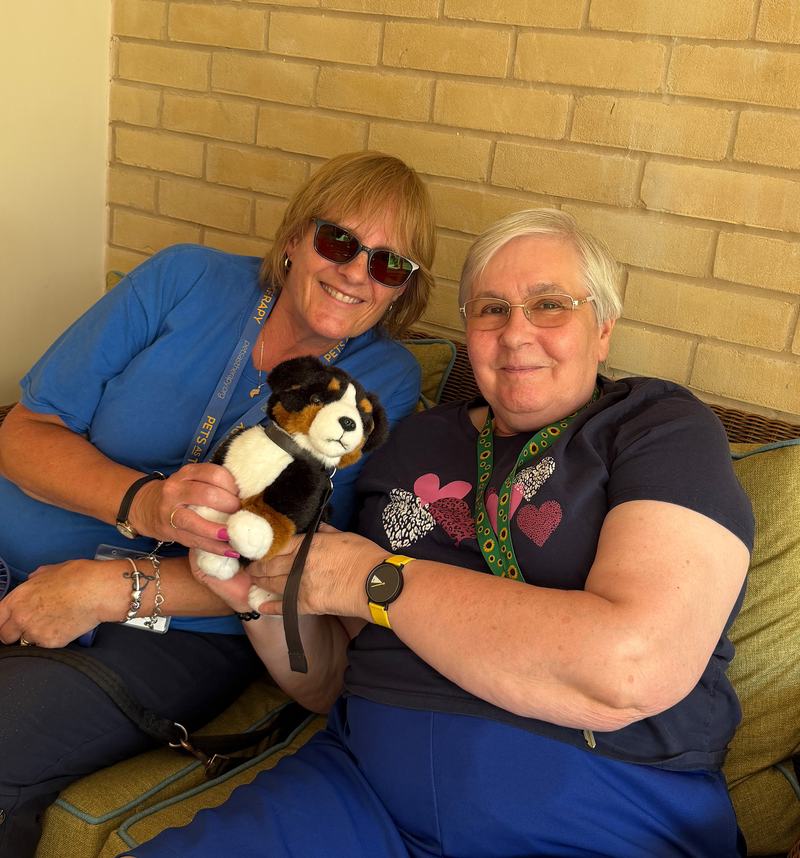 Two women are smiling and sat down together on a sofa hold a toy dog.