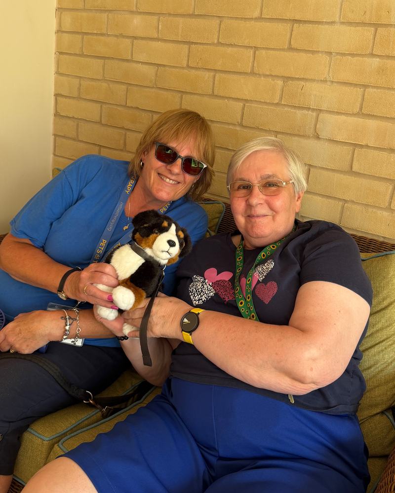 Two women are smiling and sat down together on a sofa hold a toy dog.