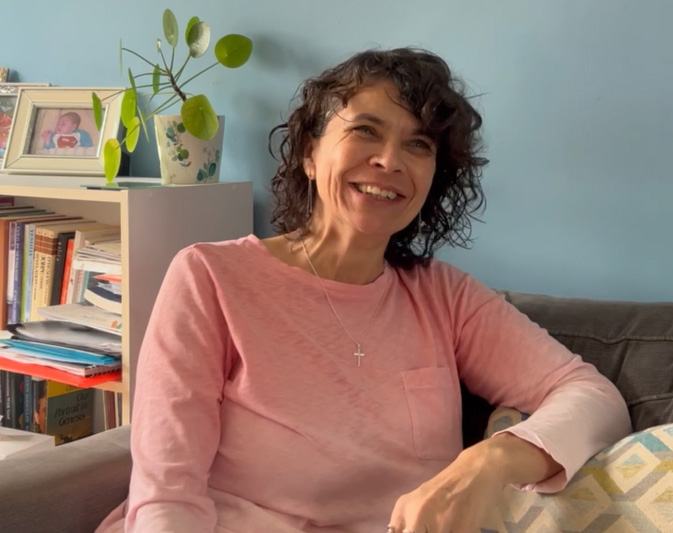 A woman with curly hair wearing a long sleeve pink top is sitting down on a sofa and smiling.