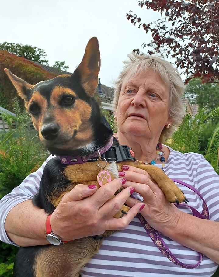 A woman holds a small black and tan dog with pointy ears. She is standing outdoors, surrounded by greenery and trees. The dog has a pink paw-shaped tag on its collar. The sky is overcast.